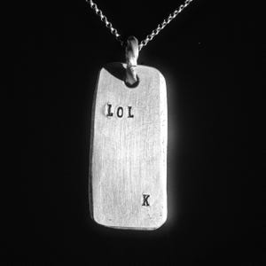 Peut inclure: Pendentif argenté en forme de plaque d'identité avec les lettres « LOL » et « K » estampillées. Il est suspendu à une fine chaîne. Le fond est noir, mettant en valeur le métal brillant.