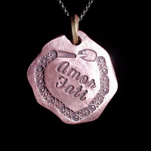 Peut inclure: Un collier pendentif de couleur cuivrée avec les mots "Amor Fati" gravés dans une police stylisée. Le pendentif a une forme unique avec une bordure décorative et est suspendu à une fine chaîne argentée.