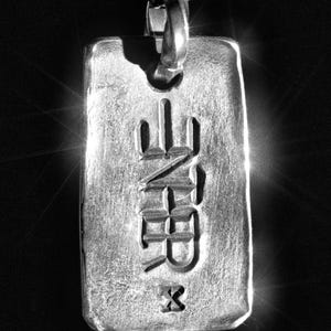Peut inclure: Pendentif rectangulaire argenté avec une chaîne. Le pendentif porte des caractères japonais gravés et un petit symbole 'X' en bas. Il est suspendu sur un fond sombre, avec la lumière se reflétant sur le métal.