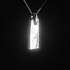 Peut inclure: Un collier pendentif argenté avec un pendentif rectangulaire texturé. Le pendentif présente un petit détail rond et lumineux près du bas. La chaîne du collier est délicate et argentée, sur fond sombre.