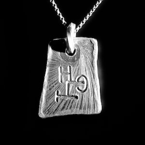 Peut inclure: Un collier avec un pendentif en argent. Le pendentif rectangulaire et texturé porte les lettres "GTH" gravées, sur un fond texturé. La chaîne du collier est également en argent et est attachée au pendentif.