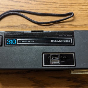 May include: A black and gray 310 BerkeyKeystone camera with a f/8 lens. The camera is made in the USA and has a black strap.
