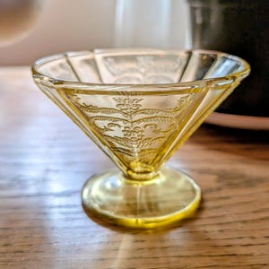 May include: A vintage yellow glass dessert dish with a decorative floral pattern. The bowl has a square top and a round base. The glass is transparent, allowing light to pass through. The dish is likely from the Art Deco era.