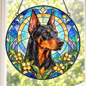 May include: A circular stained glass suncatcher featuring a Doberman Pinscher portrait in black and tan. The dog is framed by a blue and yellow geometric background with yellow flowers and green leaves at the base, suspended by a black metal chain.