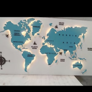 Op de afbeelding: Een turquoise wereldkaart met verlichte continenten op een witte achtergrond. De kaart bevat labels voor oceanen en continenten, een kompasroos en kleine vliegtuigpictogrammen. Een decoratief wandkunstwerk.