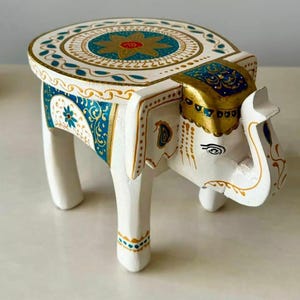 Puede incluir: Un taburete de elefante blanco pintado a mano con una parte superior redonda. El taburete presenta intrincados diseños en oro, verde azulado y azul. La cabeza y el cuerpo del elefante están detallados con patrones pintados y acentos decorativos.