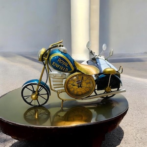 Könnte beinhalten: Ein dekoratives Motorradmodell in Blau und Gold mit dem Schriftzug "Royal Enfield" auf dem Tank. Das Modell hat eine Uhr als Vorderrad, einen goldfarbenen Sitz und Lenker. Das Motorrad steht auf einer reflektierenden Oberfläche.