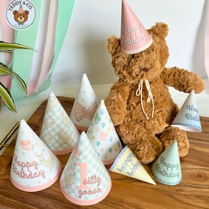 Puede incluir: Un oso de peluche marrón con un gorro de fiesta rosa con el texto "Happy Birthday", rodeado de varios gorros de fiesta en forma de cono. Los gorros presentan varios diseños, incluyendo globos, lazos y el texto "silly goose".