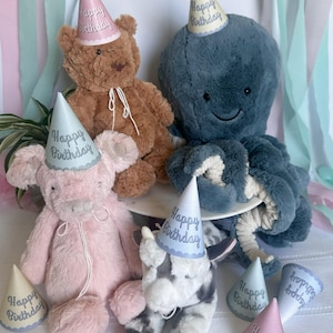 May include: A collection of plush toys, including a brown bear, a pink bunny, a blue octopus, and a black and white cow, all wearing party hats that say "Happy Birthday". The toys are arranged on a white surface with pastel-colored streamers in the background.