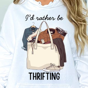 May include: White hoodie with the text "I'd rather be thrifting" and a graphic of a tote bag filled with clothes, a camera, and a shoe. The tote bag is beige, and the clothes include a denim jacket and a brown jacket.