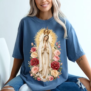 May include: A blue t-shirt featuring a religious graphic. The graphic depicts a woman in a white robe, gold crown, and halo, surrounded by roses. The design is centered on the shirt.
