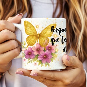 May include: White ceramic mug featuring a golden butterfly, pink and purple flowers, and the text "Porque que te d". The mug has a white handle and is held by a person.