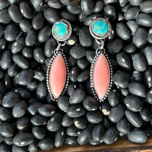 May include: A pair of silver earrings with turquoise and pink coral stones. The earrings have a teardrop shape and are set against a background of black beans.