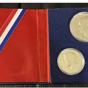May include: A United States Bicentennial Silver Proof Set in a red, white, and blue presentation case. The case opens to reveal three silver coins: a half dollar, a quarter, and a smaller coin, all displayed on a blue background.