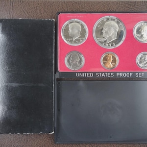 May include: A 1973 United States Proof Set in a black case. The set includes six coins displayed on a red background. The case is open, revealing the coins, with the words "United States Proof Set" printed on the case.