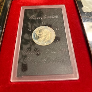 May include: A silver Eisenhower dollar coin in a protective case. The coin features a profile of President Eisenhower and the text "In God We Trust." The case is set in a red velvet box, with the words "Eisenhower" and "United States Peace Dollar" etched on the black background.