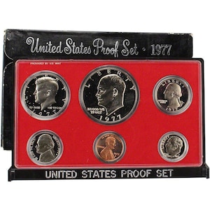 May include: A 1977 United States Proof Set in a black display case. The set includes six coins of various denominations, including a silver dollar, half dollar, quarter, dime, nickel, and penny. The coins are displayed on a red background.