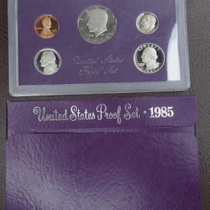 May include: A 1985 United States Proof Set in a clear plastic case, featuring various coins. The set includes a copper penny, silver coins, and a half-dollar coin. The purple case is labeled "United States Proof Set - 1985".