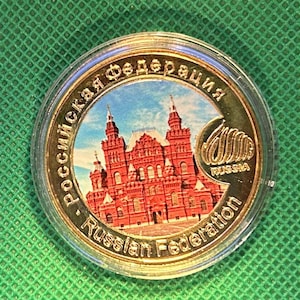May include: A gold-colored coin featuring a red building with multiple towers and windows, set against a blue sky. The coin is inscribed with the words "Russian Federation" in English and Cyrillic script, and the word "Russia".