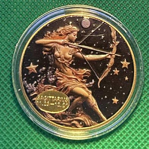 May include: A gold-toned coin featuring a woman with a bow and arrow against a starry night sky. The coin has a clear protective case and a gold rim. The woman is depicted in a flowing gown, with a crescent moon and stars in the background.