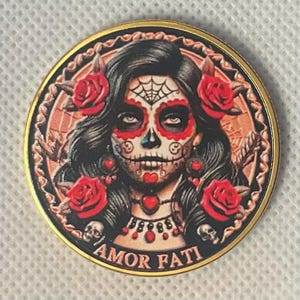 May include: A round, gold-toned coin featuring a detailed illustration of a Day of the Dead woman with sugar skull makeup, red roses, and the text "AMOR FATI". The coin has a decorative border.