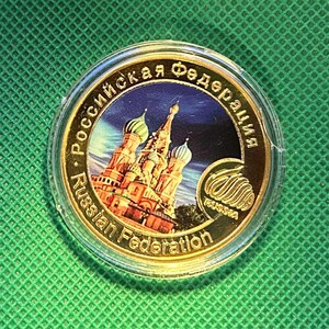 May include: A gold-colored coin featuring a detailed illustration of St. Basil's Cathedral with blue and gold domes. The coin is encased in a clear plastic holder, with text in both English and Russian. The background is a vibrant green.