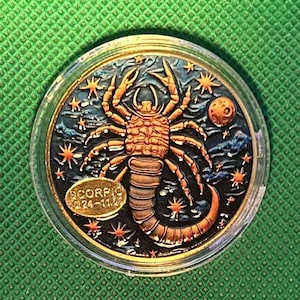 May include: A round, gold-toned coin featuring the Scorpio zodiac symbol. The coin depicts a detailed scorpion with a starry night sky background, a crescent moon, and the text "SCORPIO 10.24-11.22". The coin is encased in a clear plastic holder.