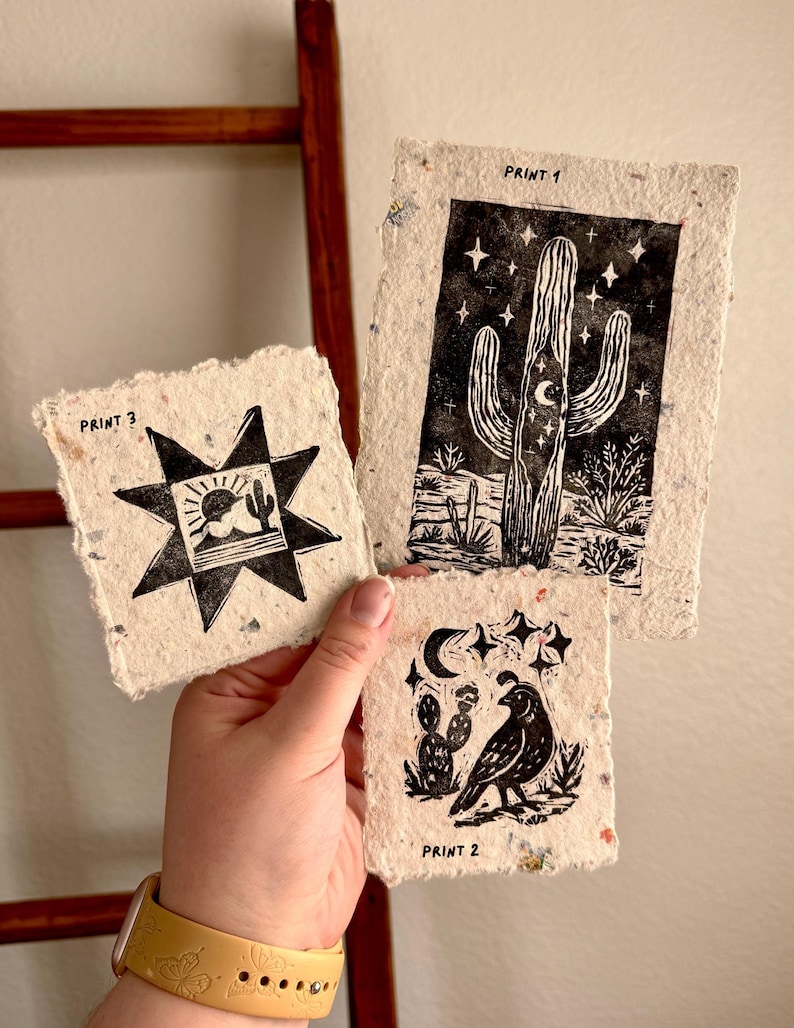 May include: Three handmade paper prints with black ink designs. Print 1 features a cactus under a crescent moon and stars. Print 2 shows a bird and cactus. Print 3 has a sun and cactus. The prints are labeled "Print 1", "Print 2", and "Print 3".