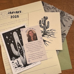 May include: A collection of stationery items featuring cactus illustrations and the text "January 2026". Includes a card with artist information and a linocut print of a cactus. The items are on a wooden surface.