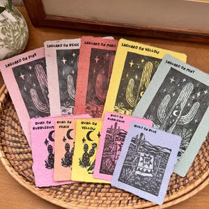 May include: A collection of colorful art prints featuring desert-themed illustrations. The prints showcase cacti, stars, and a quilt design, with text such as "Saguaro on Pink" and "Quilt on Blue." The prints are arranged in a woven basket.