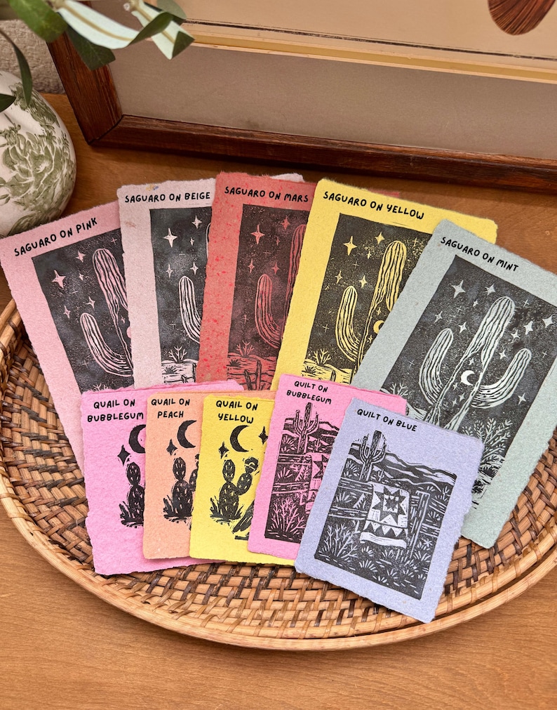 May include: A collection of colorful art prints featuring desert-themed illustrations. The prints showcase cacti, stars, and a quilt design, with text such as "Saguaro on Pink" and "Quilt on Blue." The prints are arranged in a woven basket.