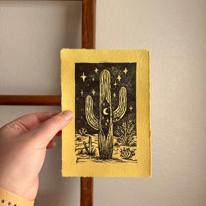 May include: A black and white linocut print of a cactus with a crescent moon and stars on a yellow background. The print depicts a desert landscape with various cacti and desert plants. The artwork is on textured paper.