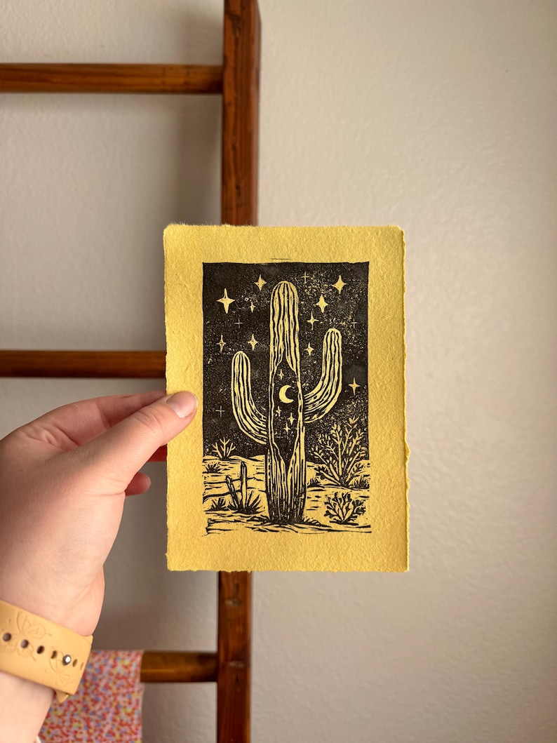 May include: A black and white linocut print of a cactus with a crescent moon and stars on a yellow background. The print depicts a desert landscape with various cacti and desert plants. The artwork is on textured paper.