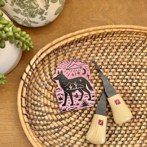 May include: A pink and black coyote design on a small rectangular block, with two wood carving tools on a woven tray. The tools have wooden handles and metal blades. The tray is brown and oval-shaped.