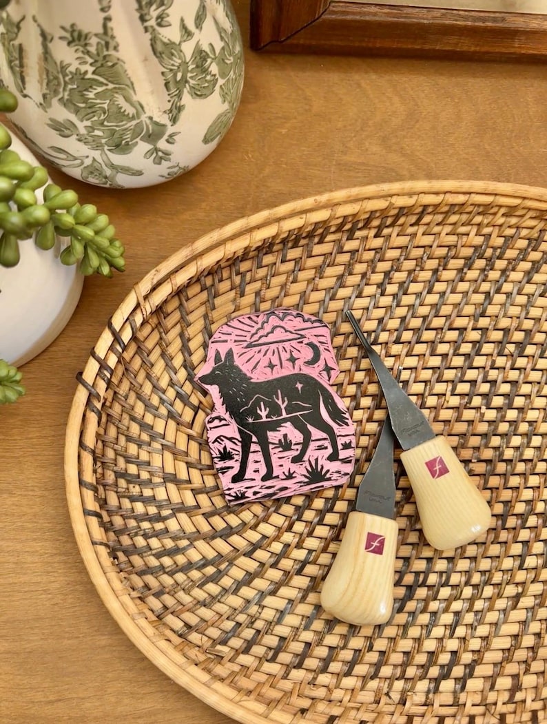 May include: A pink and black coyote design on a small rectangular block, with two wood carving tools on a woven tray. The tools have wooden handles and metal blades. The tray is brown and oval-shaped.