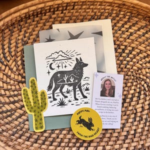 May include: A collection of desert-themed stationery items, including a cactus sticker, a card with a coyote illustration, and a round sticker with a rabbit design. The items are arranged on a woven basket.