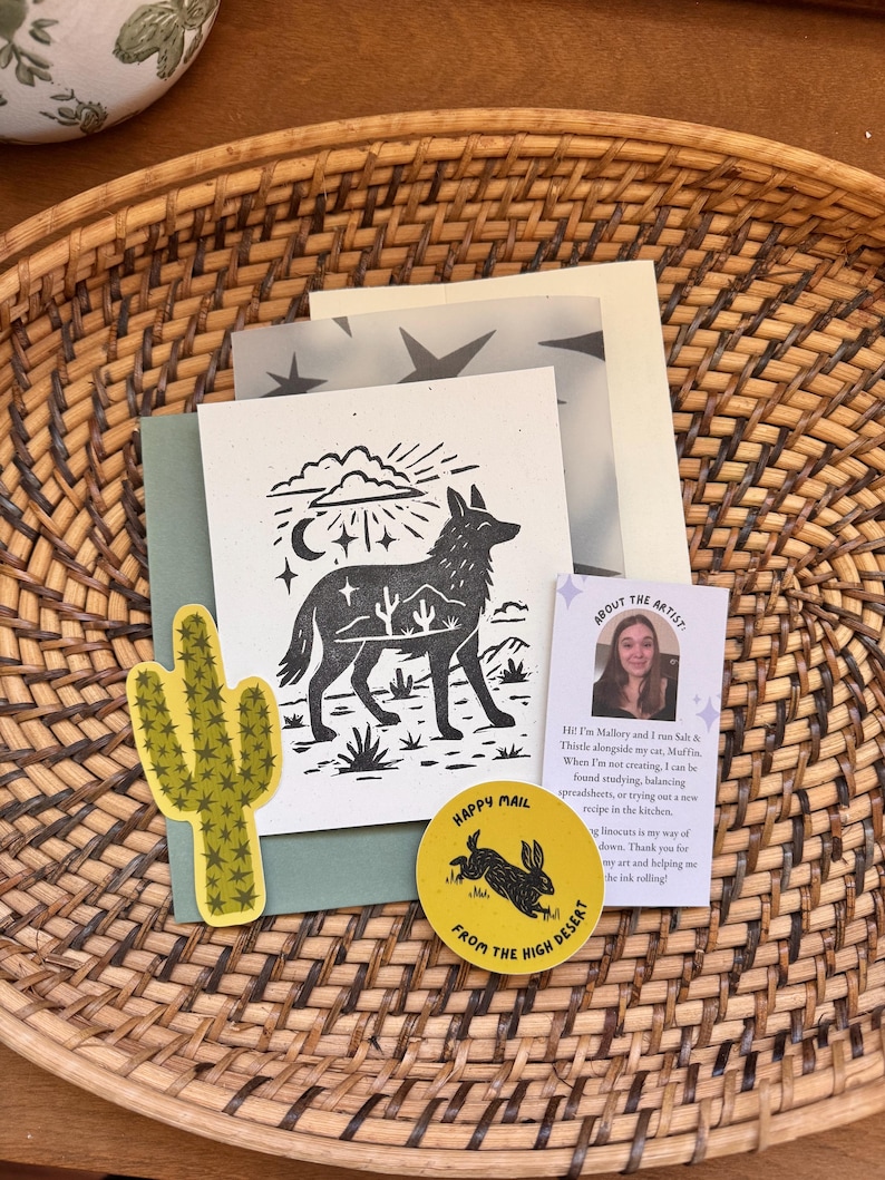 May include: A collection of desert-themed stationery items, including a cactus sticker, a card with a coyote illustration, and a round sticker with a rabbit design. The items are arranged on a woven basket.