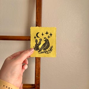 May include: A small, yellow artwork featuring a black bird, cactus, moon, and stars. The linocut print is held in a hand, with a wooden ladder in the background. The artwork has a rustic, handmade aesthetic.