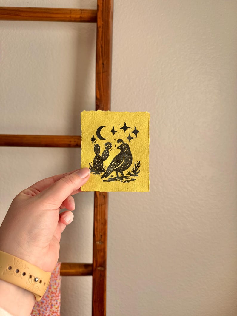 May include: A small, yellow artwork featuring a black bird, cactus, moon, and stars. The linocut print is held in a hand, with a wooden ladder in the background. The artwork has a rustic, handmade aesthetic.