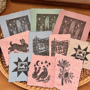 May include: A collection of handmade paper art prints featuring desert-themed linocut designs. Prints include cacti, rabbits, and starbursts in shades of pink, blue, and green. The prints are arranged in a woven basket.