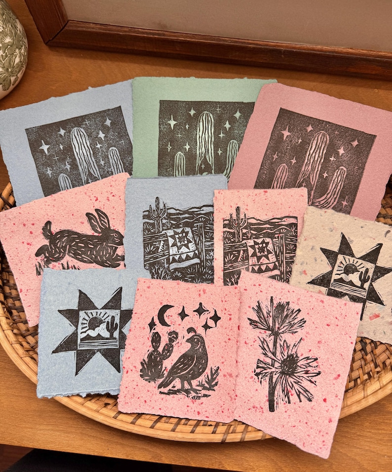 May include: A collection of handmade paper art prints featuring desert-themed linocut designs. Prints include cacti, rabbits, and starbursts in shades of pink, blue, and green. The prints are arranged in a woven basket.