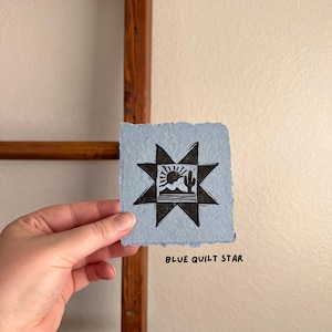 May include: A blue paper with a black star design and a desert landscape illustration. The text "BLUE QUILT STAR" is printed in black below the image. The paper has a rough, textured edge.