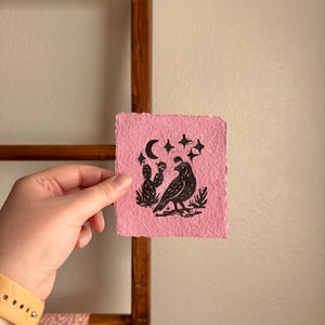May include: A pink, square art print featuring a black illustration of a bird, cactus, moon, and stars. The print is held in a hand, with a wooden ladder and patterned fabric in the background.