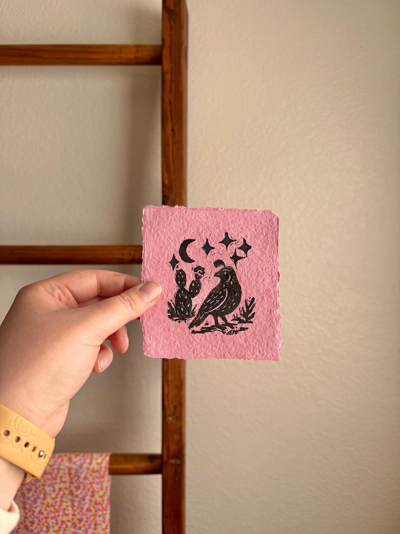 May include: A pink, square art print featuring a black illustration of a bird, cactus, moon, and stars. The print is held in a hand, with a wooden ladder and patterned fabric in the background.