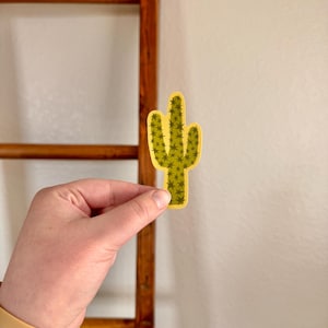 May include: A yellow and green cactus sticker with a detailed design. The sticker is held in front of a wooden ladder and a white wall. The cactus has a detailed pattern of small green spikes.