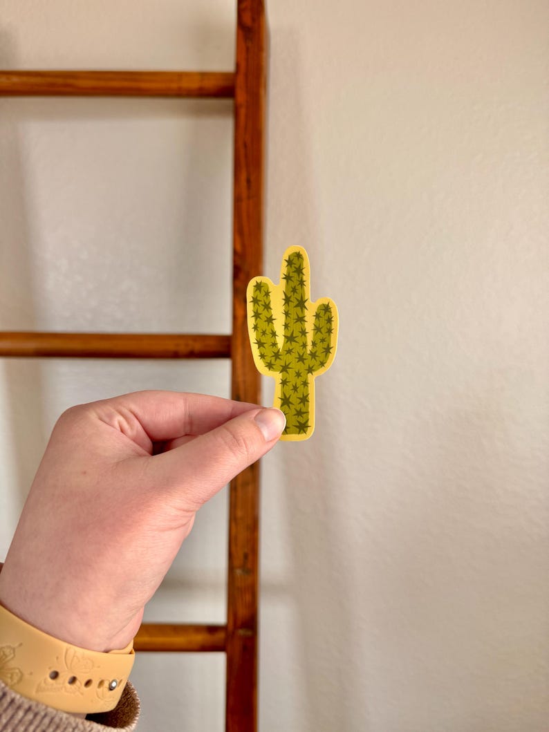 May include: A yellow and green cactus sticker with a detailed design. The sticker is held in front of a wooden ladder and a white wall. The cactus has a detailed pattern of small green spikes.