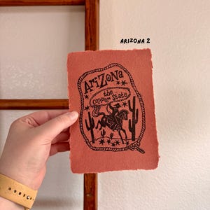 May include: A hand holds a textured, reddish-orange print with a black illustration of a cowboy on a horse, cacti, and the text "Arizona the Copper State." The print is labeled "Arizona 2."