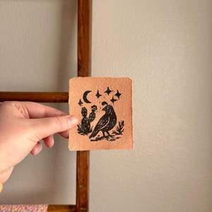 May include: A small, square, handmade paper art piece with a black linocut print. The print features a bird, cactus, moon, and stars. The paper is a light brown color, and the print has a rustic, handmade aesthetic.