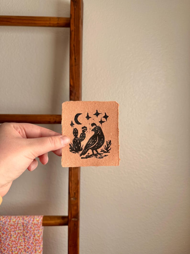 May include: A small, square, handmade paper art piece with a black linocut print. The print features a bird, cactus, moon, and stars. The paper is a light brown color, and the print has a rustic, handmade aesthetic.