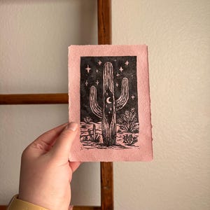 May include: A pink print featuring a black cactus design with a crescent moon and stars. The artwork is a linocut print, showcasing a desert landscape. The print is held against a wooden ladder.